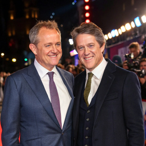 Hugh Bonneville and Hugh Grant at a film premiere. Both in tailored suits. Bright lights and crowd in the background.