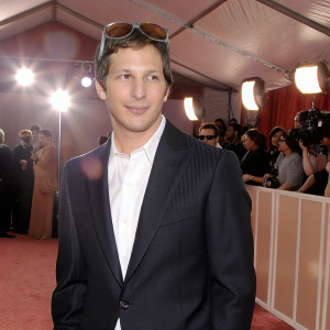 Andy Samberg, wearing a black suit and white shirt, sunglasses perched atop his head. Amidst crowd at Grammy Awards. Studio audience and bright lights.