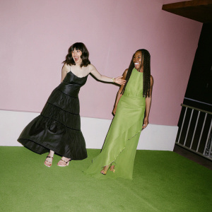 Carly Henderson and Erinn Westbrook at the MTVU Woodie Awards. Both are smiling. Purple backdrop and glossy black floor. Carly in black dress with accents, wavy brown hair. Erinn in bright yellow dress, straight black hair.