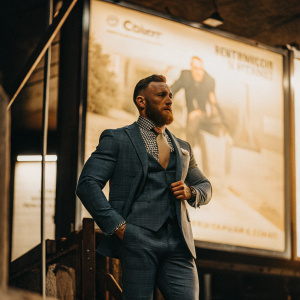 Conor McGregor, bearded and wearing a blue suit with a checked shirt and tie, poses confidently in front of a large billboard advertising his upcoming fight.
