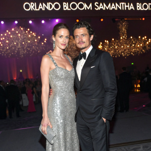Orlando Bloom and Samantha Bloom at an exclusive gala. She's wearing a dazzling silver gown, he in a sleek black tuxedo. Behind them, a dimly lit stage with sparkling chandeliers and a large banner declaring the event's theme.