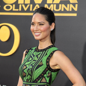 Olivia Munn, Asian-American actress, smiling confidently at a glitzy awards show. She's dressed in a vibrant green and black sleeveless dress with intricate patterns. The backdrop features bold yellow logo text elements.