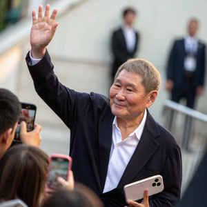 A blurry background photo of Takeshi Kitano waving, captured by an enthusiastic fan holding a large phone.