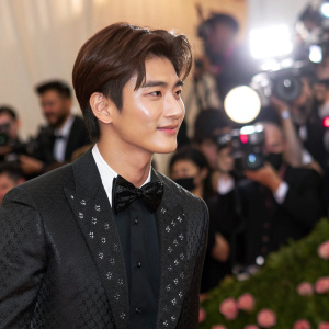 Park Hyung Sik at the Met Gala. Wearing a black suit with a bow tie. Surrounded by paparazzi, flashing camera lights