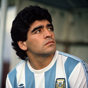 Diego Maradona in a candid shot. Dark hair, intense gaze. Wearing an AFA jersey. Studio setting with blurred background