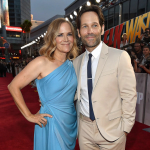 Paul Rudd and Julie Yaeger at the Ant-Man and the Wasp premiere. Both are dressed in formal attire. She wears a blue, sleeveless dress with a draped silhouette, he sports a beige suit, white shirt, and white tie. Flashing lights and fans in the background.