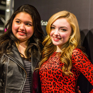 Charli Barcena and Peyton List, both with smiles, at a promotional event. Charli in a black leather jacket, Peyton in a leopard print top. They pose against a plain black backdrop with corporate logo.