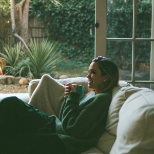 Jessica Alba in a cozy home setting. Wearing a casual green sweater. Sitting on a white sofa. Background features an indoor garden with lush greenery.
