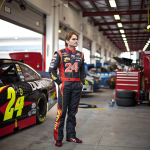 Jeff Gordon, the NASCAR driver, stands beside his race car. He wears number 24. The background is a bustling garage area with various mechanics and vehicles.