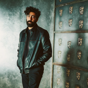 Ray Panthaki, British actor, posing at BAFTA awards. Bearded, curly hair, black leather jacket, standing alone against backdrop with BAFTA logos.
