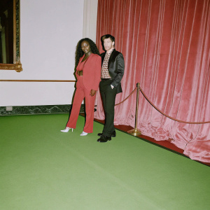 Beverley Knight and James O'Keefe at a glamorous award show. Rich red velvet curtain backdrop. Beverley in a bold red ensemble. James in a sleek dark blazer and patterned shirt.
