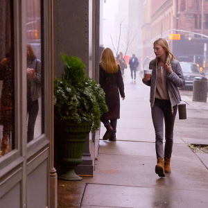 Whitney Port, blonde woman in casual attire, walking past a trendy coffee shop. Green potted plant beside her. Another blonde woman walking away from the camera on a busy city street.