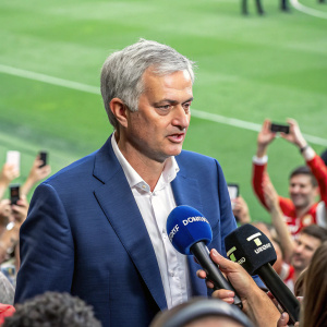 Jose Mourinho, wearing a blue suit, addressing the media. Background filled with green grass and cheering fans.