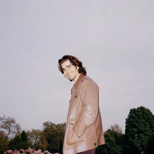 Eric Cantona, smiling warmly. Wearing a brown leather jacket. Standing in a sunny park.