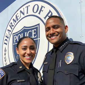 Diara Cruz and Patrick Espeut, two police officers, smiling for a department photo. They wear dark uniforms and badges. A police station wall with a large department logo stands behind them.