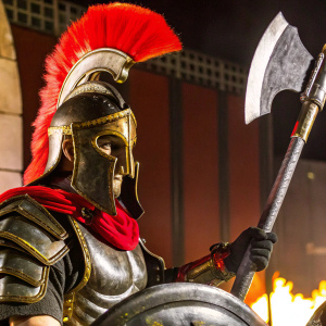 In the fiery realm of Sparta: War of Empires, a Spartan warrior stands vigilant. Helmeted with a red plume, clad in a red scarf, he wields a large, ornate battle axe. Dark red background.