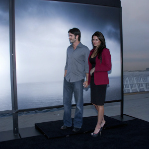 Brandon Beemer and Nadia Bjorlin at a film festival premiere. They are dressed in formal attire. Brandon in gray shirt and blue jeans, Nadia in red blazer and black dress. Black carpeted area with movie poster backdrop.