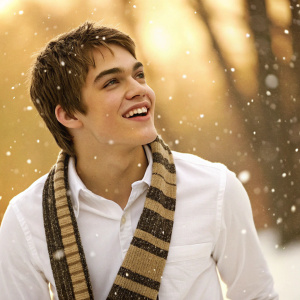 Cheerful Dylan Sprayberry, young actor, in a cozy winter scene. White shirt and striped scarf. Warm, golden background.