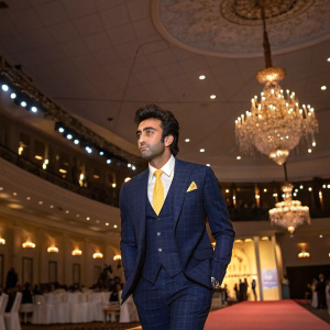 Ranbir Kapoor at the GQ Men of the Year Awards. Wearing a navy blue suit with a yellow tie. The venue is a grand hall with chandeliers.