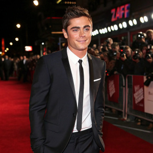 Zac Efron at award show. Stylish black suit, white shirt, black tie. Red carpet ambiance. Crowds and cameras in the background.