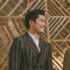 Henry Golding at the premiere event. He's smiling, wearing a dark suit with a white shirt underneath. The background is a shiny, metallic design.