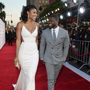Kevin Hart and Eniko Parrish walking on a red carpet event. Kevin Hart in a grey suit. Eniko Parrish in a white gown.
