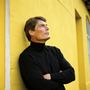 Christopher Reeve, actor, stands against a bright yellow wall with his arms crossed, wearing a black turtleneck. Intense gaze