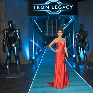 Serinda Swan, dressed in a vibrant red gown, posing at the opening night of Tron Legacy. Surrounded by sleek, metallic robot figures. Futuristic set design with large 'Tron Legacy' logo