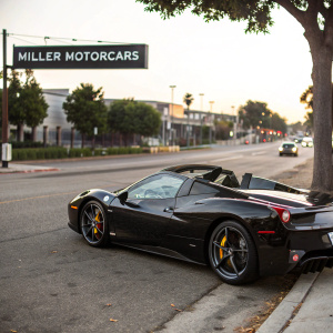Luxury black Ferrari 458 Spider parked in a street scene with 'Miller Motorcars' sign in background.