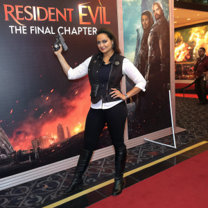 Sonakshi Sinha promoting Resident Evil: The Final Chapter. Wears black vest, white shirt, black leggings, and boots. Stands in front of large movie advertisement. Poses confidently with prop gun.
