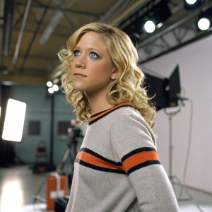 Brittany Snow as Meg Pryor on set of 'American Dreams'. Blonde curls, gray sweater with orange and black accents. Studio environment with lighting equipment, artificial illumination.