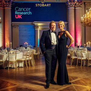 Michael Flatley in a tuxedo and Niamh O'Brien in a black dress, posing for a charity event photo. The backdrop is filled with the logos of Cancer Research UK and Stobart. The event is held in a grand ballroom with a lavish chandelier and tables set for a gala dinner.