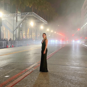 Ronda Rousey at film festival. Wearing a black dress, standing alone. Nighttime street with bright lights in background.