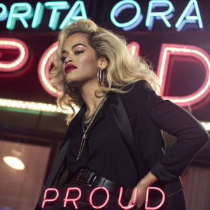 Rita Ora, blonde hair, in a stylish black outfit. Promoting her single 'Proud'. Shot against a dynamic, neon-lit background.