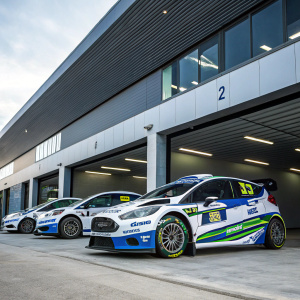 Several Ford Fiesta R5 rally cars, bedecked with sponsor logos, parked at a sleek modern garage. Each car displays a vibrant color scheme, predominantly white and blue with green highlights. Emphasizing their racing capabilities and dynamic design.