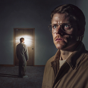 Promotional photo for 'Manhunt: Unabomber'. The gentleman, wearing glasses, stands alone in an empty, dimly lit room with a single spotlight.
