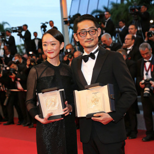 Zhou Xun and Masahiro Motoki accepting awards at Cannes Film Festival. They are in formal attire. Red carpet, paparazzi flashing cameras.