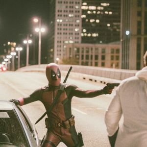 Comic panel featuring Wade Wilson aka Deadpool, standing in a heroic pose. Background: city skyline at night. Colleague in fighting stance beside him.
