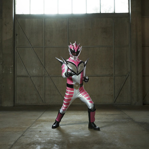 Kamen Rider Ryuki, depicted in a dynamic pose, adorned with a fierce mask design featuring sharp edges and jagged patterns. The vibrant colors of pink and gray contrast against a dark, dramatic background.