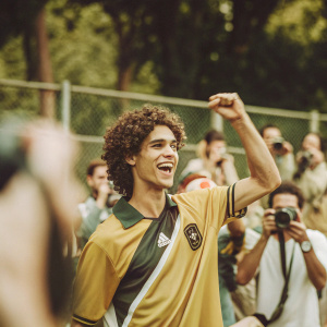 Raphael Varane, sporting curly hair and wearing a jersey with team crest, celebrates on the sidelines. A sea of photographers in the background.
