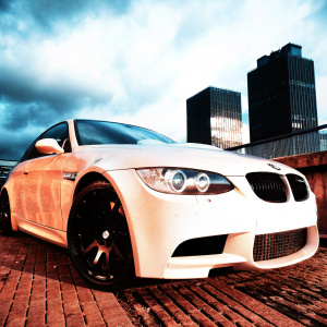 The BMW M3, gleaming in the twilight, stands out against the grey urban skyline. Dark clouds loom overhead, casting shadows that dance across its polished exterior. Immaculate black wheels and sleek aerodynamic design emphasize its power and elegance.