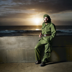 Che Guevara depicted in vibrant color palette. Wearing green military fatigues and standing against a sunset backdrop. Waves crashing behind him.