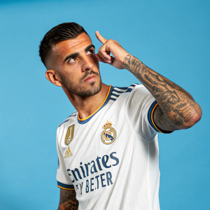 Dani Ceballos, Hispanic soccer player with a sharp pose. Close-cropped hair and tattoos on arms. Wearing Real Madrid jersey. Bright blue background.