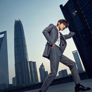 A promotional image for Kim Taehyung, featuring him in a stylish suit with a dynamic pose. Dark urban cityscape background, modern and sleek design.