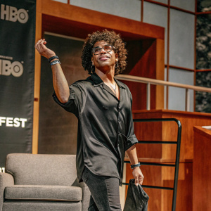 Christopher Gray at the Outfest Los Angeles LGBTQ Film Festival. Standing confidently with curly hair. Wearing a black shirt. The backdrop features HBO branding.
