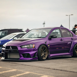 John Mazo's custom purple Mitsubishi Lancer Evo JD Edition. Shiny paintwork and sporty design. Blurred background of parked vehicles.
