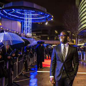 Tinie Tempah, dressed in a sleek black suit and vibrant white shirt, with a black and white striped tie, graces the 2011 Brit Awards red carpet. His charming smile radiates confidence as he poses for the cameras.