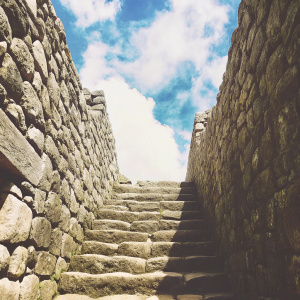Ancient stone steps ascending at Machu Picchu, Peru. Stone walls crafted from irregularly shaped stones, weathered by time. Vibrant blue sky with fluffy clouds. Ground level view looking upwards along the path towards the sky.