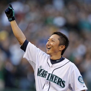 Munenori Kawasaki, a cheerful Asian baseball player, in a white Seattle Mariners jersey, celebrating with raised arm. Crowd blurred in background.