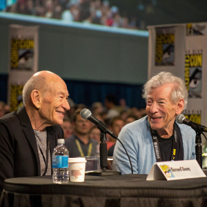 Patrick Stewart and Ian McKellen during an interview at Comic-Con. Seated at a round table with microphones and a water bottle. Convention crowd in the background.